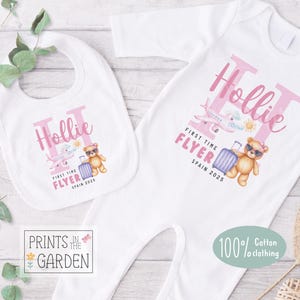May include: A white baby bib and sleepsuit set. Both items display a pink design with the name "Hollie", a teddy bear with sunglasses, a suitcase, and an airplane. The text "FIRST TIME FLYER SPAIN 2025" is also included.