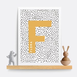 Puede incluir: Una impresi&oacute;n enmarcada en blanco con un fondo de lunares blanco y negro. Una gran letra F amarilla se encuentra en el centro de la impresi&oacute;n. El nombre "FLORA" est&aacute; escrito verticalmente en el lado izquierdo de la letra F.