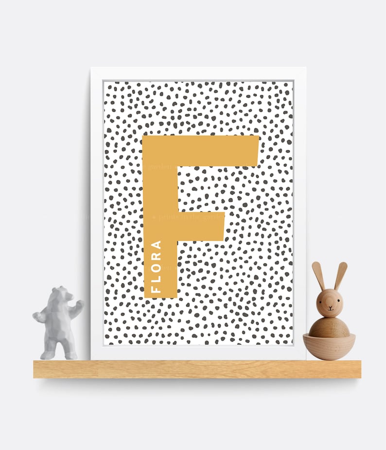 Puede incluir: Una impresi&oacute;n enmarcada en blanco con un fondo de lunares blanco y negro. Una gran letra F amarilla se encuentra en el centro de la impresi&oacute;n. El nombre "FLORA" est&aacute; escrito verticalmente en el lado izquierdo de la letra F.