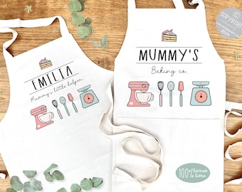 May include: Two white aprons with pink and blue baking themed designs. The smaller apron says "EMILIA Mommy's little helper." The larger apron says "MUMMY'S Baking co."
