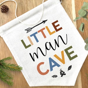 Op de afbeelding: Een witte canvasbanner met de tekst "LITTLE man CAVE" in kleurrijke letters. De letters zijn in verschillende kleuren, waaronder groen, oranje, geel en blauw. Boven het woord "LITTLE" staat een kleine pijl en onder het woord "CAVE" een klein kampvuurontwerp.