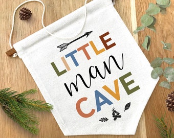 May include: A white canvas banner with the text "LITTLE man CAVE" in colorful letters. The letters are in different colors, including green, orange, yellow, and blue. There is a small arrow above the word "LITTLE" and a small campfire design below the word "CAVE".