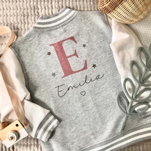 May include: Gray and white baseball style jacket with a pink letter E and the name Emilia on the back. The jacket has white sleeves with gray stripes on the cuffs.