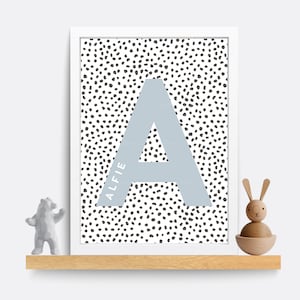 Puede incluir: Una impresi&oacute;n enmarcada en blanco con una letra A azul claro sobre un fondo de lunares negros y blancos. El nombre Alfie est&aacute; escrito verticalmente en el lado izquierdo de la letra A.