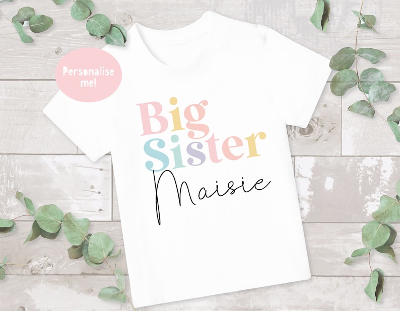 May include: White t-shirt with the text "Big Sister" in colorful letters and the name "Maisie" written in cursive below.