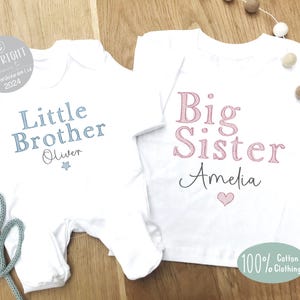 May include: Two white baby onesies, one with blue text that says "Little Brother Oliver" and a blue star, and the other with pink text that says "Big Sister Amelia" and a pink heart.