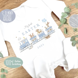 May include: A white baby grow with a blue train design featuring a bunny, lamb, and other Easter-themed characters. The grow has the text "My first Easter" and "2025" printed on it. The grow is personalised with the name "Charlie".