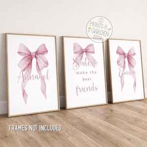 May include: Three vertical wall art prints in light wood frames. Each print features a watercolour-style pink ribbon bow. The left print says Annabel, the centre says Sisters make the best friends, and the right says Emilia. Text reads FRAMES NOT INCLUDED.