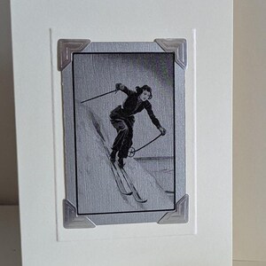May include: A vintage black and white photo of a person skiing down a snowy slope. The photo is framed with a silver border and is on a white card with a silver embossed border. The card has a logo in the bottom right corner that says "Fuzzy Mouse".