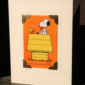 Peut inclure: Carte de v&oelig;ux avec Snoopy et Woodstock jouant aux cartes sur le niche de Snoopy. L'illustration est sur un fond orange et encadr&eacute;e de coins dor&eacute;s. La carte est sur un fond blanc.