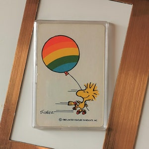 May include: A vintage Snoopy card featuring Woodstock holding a rainbow balloon, with the text "SCHULZ" and "© 1965 UNITED FEATURE SYNDICATE, INC." in a clear plastic case, set against a copper-colored frame.