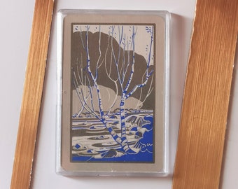 Op de afbeelding: Een kaart in vintage stijl met een blauwe en witte illustratie van een boom met kale takken in een besneeuwd landschap. De kaart is ingelijst in een gouden lijst.