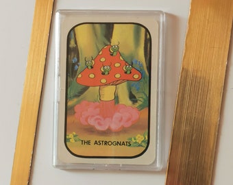 Op de afbeelding: Een vintage verzamelkaart met een cartoonillustratie van een rode en gele paddenstoel met groene en gele beestjes erop. De kaart heeft een gouden rand en de tekst "THE ASTROGNATS" onderaan.