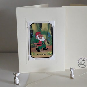 May include: A white card with a picture of a gnome in a green suit and red hat walking in a forest. The card has a white border and the text "The Gnome" at the bottom. The card is on a white surface with a small white mouse figurine.
