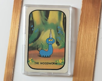 Op de afbeelding: Een ingelijste vintage ruilkaart met een cartoon blauw Woodworm-personage met een eenhoornhoorn. De kaart staat tegen een achtergrond van een boom en bloemen, in een doorzichtige plastic hoes en ingelijst in een goudkleurig metalen frame.