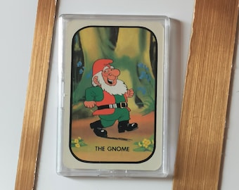 Op de afbeelding: Een ingelijste kaart met een cartoon dwerg in groene en rode kleding, met de tekst "THE GNOME" onderaan. De kaart is beschermd door een transparante plastic hoes en geplaatst in een goudkleurig rechthoekig frame.
