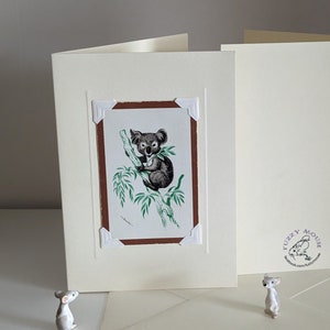 May include: A blank card with a brown frame and a picture of a koala bear inside. The card is on a white surface with two small white mice figurines.