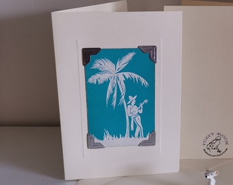Op de afbeelding: Een witte kaart met een turquoiseblauwe afbeelding van een palmboom en een persoon die ukulele speelt. De afbeelding is omlijst met zilveren hoeken. De kaart heeft een witte envelop en een klein wit muisbeeldje.