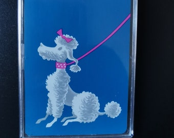 Op de afbeelding: Een witte poedel met een roze strik en een halsband op een blauwe achtergrond. De poedel zit en kijkt naar links. Een roze riem is aan de halsband bevestigd en loopt naar rechts uit het beeld.