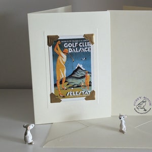 May include: A white card with a gold border and a vintage poster of a woman playing golf. The poster is for the Golf Club Dalsace in Sélestat, France. The card has a Fuzzy Mouse logo on the back.