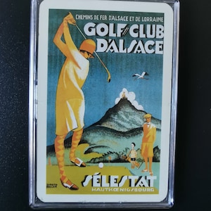 May include: A vintage travel poster advertising the Golf Club D'Alsace in Sélestat, Haut-Koenigsbourg. The poster features a woman in a yellow dress and white hat playing golf, with a mountain and a bird in the background.
