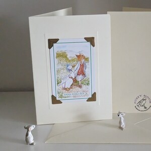 May include: A white card with a gold border and a picture of a fox and a duck from the book "Jemima Puddle-Duck" by Beatrix Potter. The card is on a white surface with two small white mice figurines.