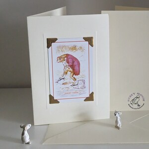 May include: A white card with a gold border and a picture of a frog wearing a red jacket and holding a shovel. The card has the text "Jeremy Fisher" on it. The card is sitting on a white surface.