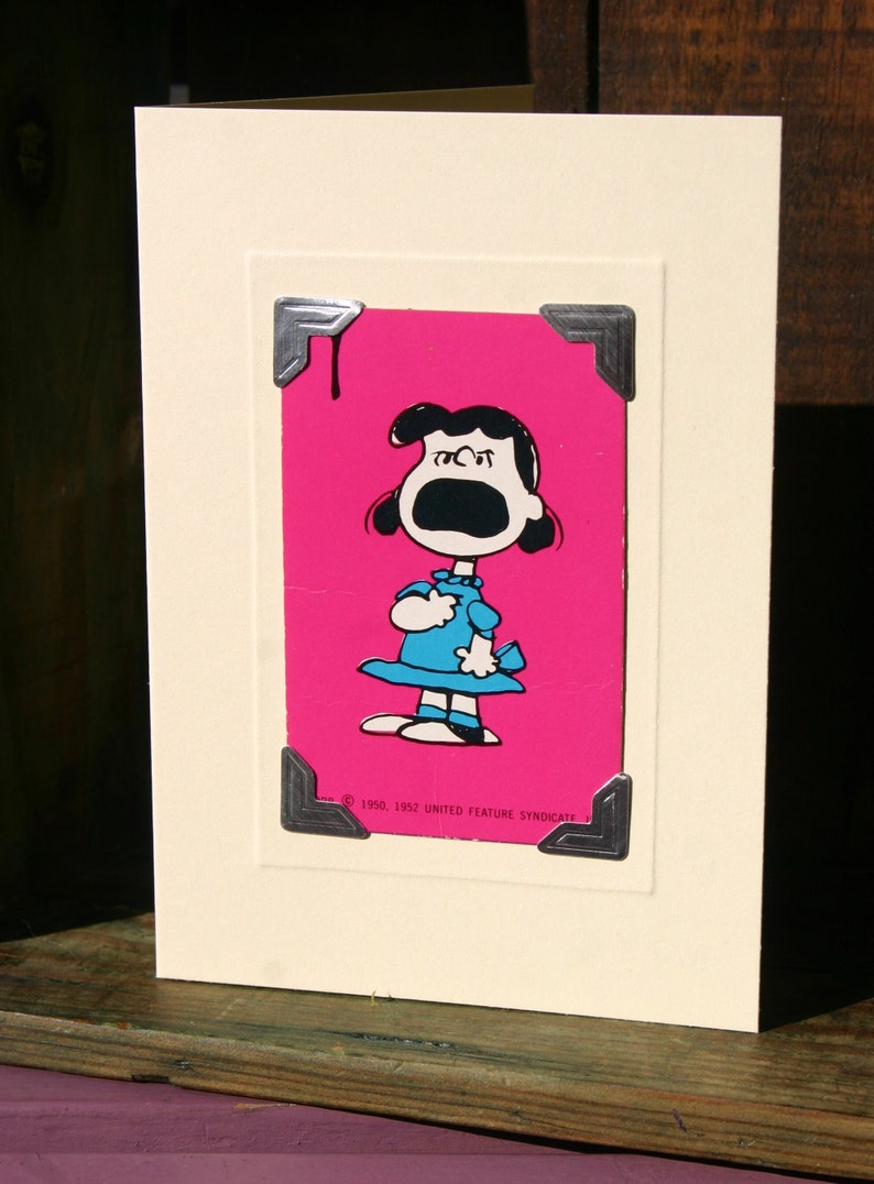 May include: A vintage card with a pink background and a black and white illustration of a girl with her mouth open, as if she is yelling. The card has a silver frame with decorative corners and the text "&copy; 1950, 1952 UNITED FEATURE SYNDICATE".