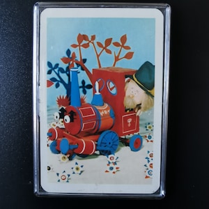 May include: A red and blue toy train with a white and red striped caboose. A bearded man in a brown hat is peeking out of the caboose window. The train is on a white background with white flowers.