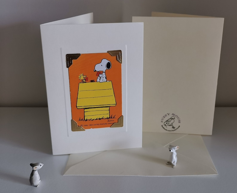Peut inclure: Carte de v&oelig;ux blanche avec Snoopy et Woodstock sur fond orange, avec une niche jaune. La carte a des coins dor&eacute;s. Une petite figurine de souris est devant la carte.