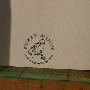 Peut inclure: Gros plan sur une carte blanche avec un tampon circulaire violet. Le tampon repr&eacute;sente une illustration de souris de dessin anim&eacute; et le texte "fuzzy mouse" et "facebook.com/fuzzymouse". La carte est pos&eacute;e sur une surface en bois.