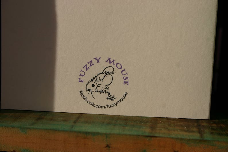 Peut inclure: Gros plan sur une carte blanche avec un tampon circulaire violet. Le tampon repr&eacute;sente une illustration de souris de dessin anim&eacute; et le texte "fuzzy mouse" et "facebook.com/fuzzymouse". La carte est pos&eacute;e sur une surface en bois.
