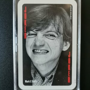 May include: A black and white playing card featuring a joker image. The card has the word "JOKER" printed vertically on both sides. The joker image is of a man with a mischievous expression. The card is encased in a clear plastic sleeve. The text "STEVE DOUBLE" and "Mark E Smith" are printed on the card.
