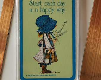 Op de afbeelding: Een vintage kaart met een meisje in een blauwe muts en een patchwork jurk, met bloemen. De kaart heeft een groene achtergrond met de tekst "Start each day in a happy way". De kaart zit in een doorzichtige plastic hoes.