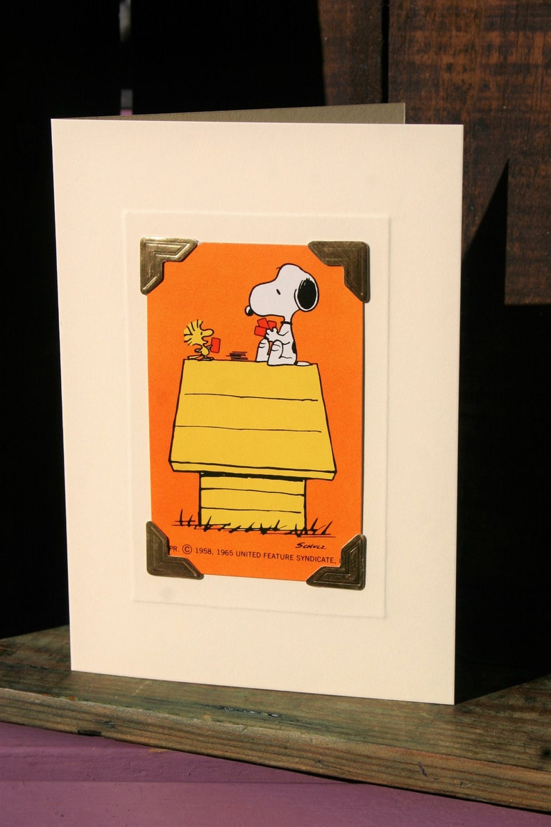 Peut inclure: Carte de v&oelig;ux avec Snoopy et Woodstock jouant aux cartes sur le niche de Snoopy. L'illustration est sur un fond orange et encadr&eacute;e de coins dor&eacute;s. La carte est sur un fond blanc.