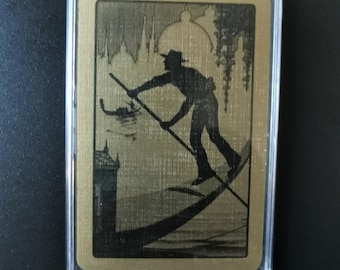 Op de afbeelding: Een goudkleurige kaart met een zwart silhouet van een gondelier die een gondel roeit voor een stadsbeeld. De kaart is ingelijst in een transparante plastic hoes.