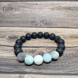 Könnte beinhalten: Ein Armband mit schwarzen Lavasteinperlen und einer Mischung aus hellblauen, weißen und grauen Edelsteinperlen. Das Armband wird auf einer Holzoberfläche präsentiert.