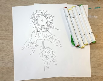May include: A black and white line drawing of a sunflower on white paper, with a stem and leaves. Next to the drawing are six white markers with colored tips, including brown, yellow, and green. The image is on a light wood surface.