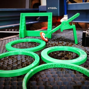 May include: Four green plastic rings with different sizes and text, "16oz Wine Tumbler", "12oz Wine Tumbler", and "16oz Pint Glass" are visible. The rings are on a black honeycomb surface. There is a green plastic tool with a lever and a small green plastic frame in the background.
