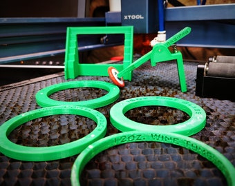 May include: Four green plastic rings with different sizes and text, "16oz Wine Tumbler", "12oz Wine Tumbler", and "16oz Pint Glass" are visible. The rings are on a black honeycomb surface. There is a green plastic tool with a lever and a small green plastic frame in the background.