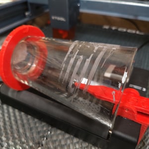 May include: A clear glass with a red plastic ring and a red plastic clamp. The glass is being held in place by the clamp on a black surface.