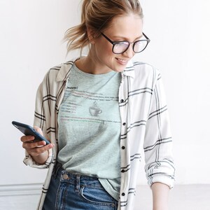 May include: A woman wearing a light blue t-shirt with a white and black plaid shirt over it. The t-shirt has a graphic of a cup of coffee and code that reads "JavaScript". She is holding a black smartphone in her left hand and looking down at it.