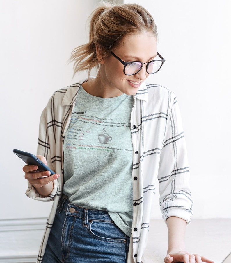 May include: A woman wearing a light blue t-shirt with a white and black plaid shirt over it. The t-shirt has a graphic of a cup of coffee and code that reads "JavaScript". She is holding a black smartphone in her left hand and looking down at it.