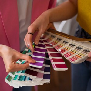 May include: Two people are looking at a color swatch book. The book is open and the pages are fanned out. The swatches are in a variety of colors, including purple, red, and green.