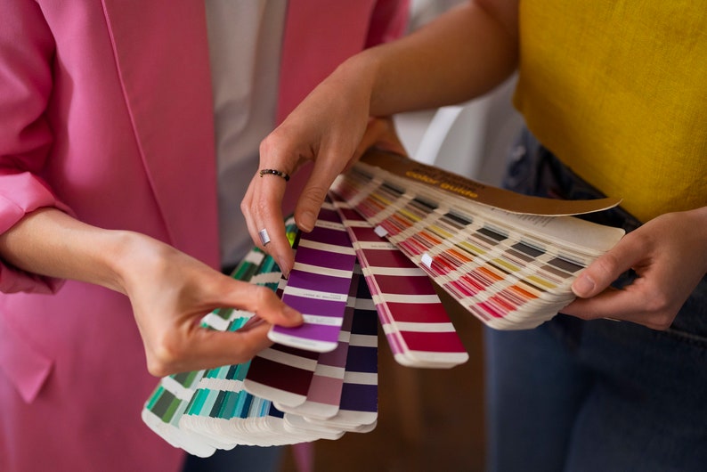 May include: Two people are looking at a color swatch book. The book is open and the pages are fanned out. The swatches are in a variety of colors, including purple, red, and green.