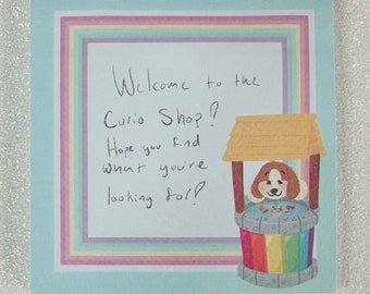 May include: A light blue card with a rainbow border. The card says "Welcome to the Curio Shop! Hope you find what you're looking for!" and features a cartoon dog in a colorful well.