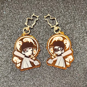 May include: Two gold-coloured keychains featuring a cartoon character holding a baseball bat. The character is wearing a baseball cap and a white and black striped shirt. The background is filled with small white ghosts.