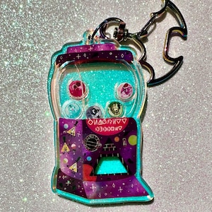 May include: A clear acrylic keychain shaped like a gumball machine, featuring a teal glitter interior and various character designs. The keychain has a silver clasp and a decorative, iridescent charm. The text "DANGER" is visible.