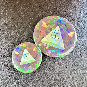 May include: Two holographic pinback buttons featuring a cartoon triangle with one eye. The larger button has a colorful background with a holographic effect. The smaller button has a similar design with a more muted background.