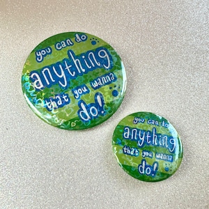 May include: Two green and blue round buttons with the text "you can do anything that you wanna do!" in blue. The larger button has a paw print and hearts on it.